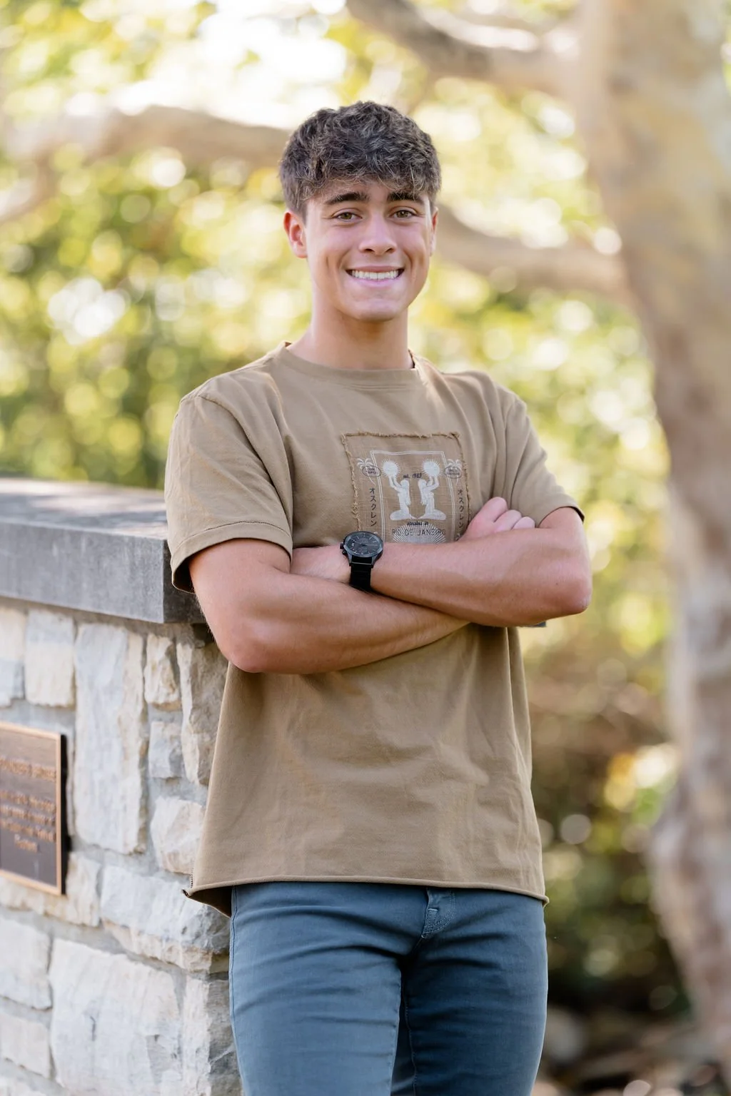 A young man with dark hair, smiling, standing outdoors with arms crossed, wearing a tan t-shirt and a black watch, next to a stone wall with a plaque, with trees and sunlight in the background.