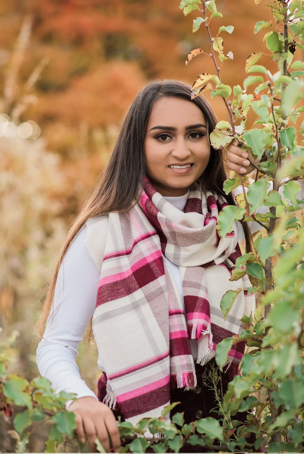 A woman with long dark hair wearing a white shirt and a red, beige, and pink plaid scarf is standing among fall foliage, holding a leafy branch and smiling.