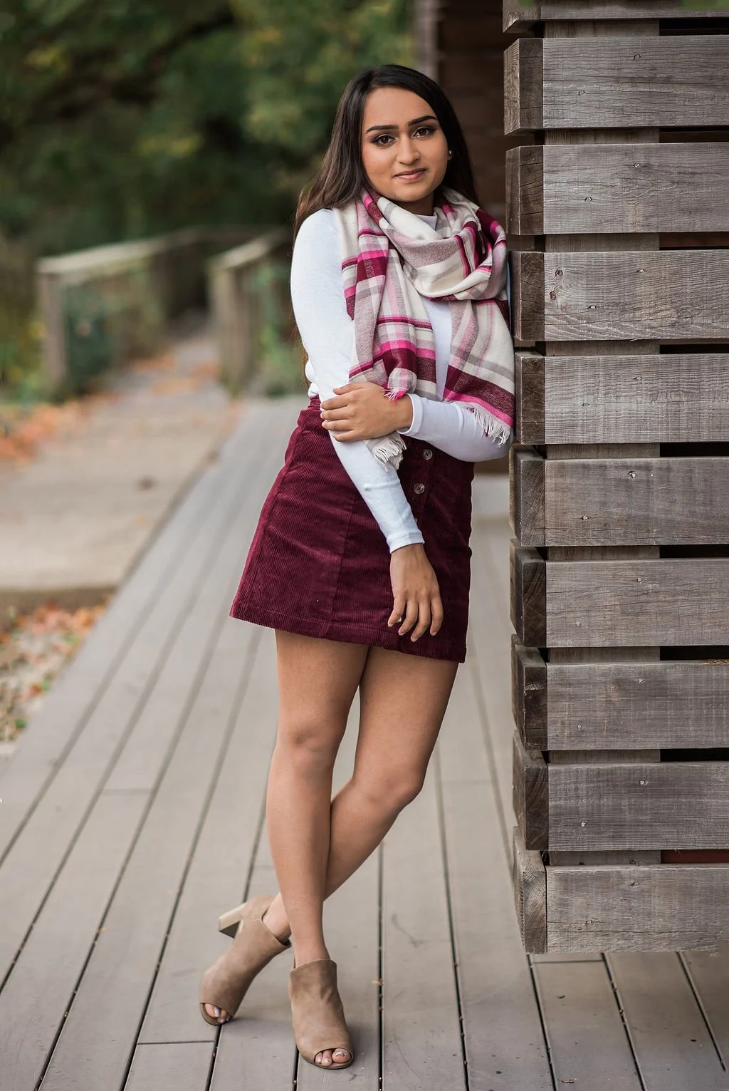 A woman in a white long-sleeve shirt, burgundy skirt, beige heels, and a plaid scarf stands on a wooden pathway outdoors, partially hidden behind a wooden structure, with a blurred background of trees and fall leaves.