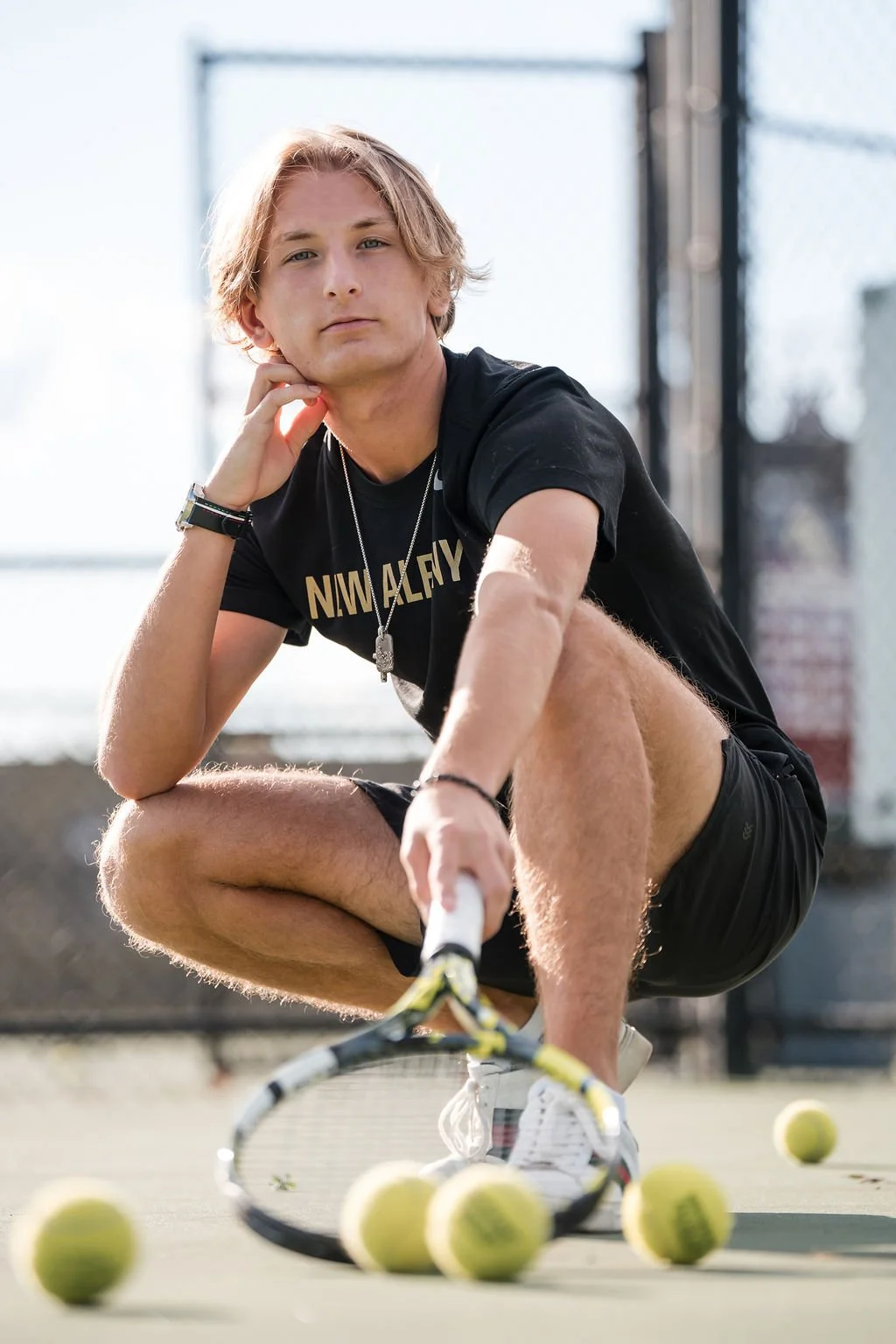 A young male tennis player squatting on an outdoor tennis court, surrounded by tennis balls, holding a tennis racket, wearing a black t-shirt and shorts, with a serious expression.