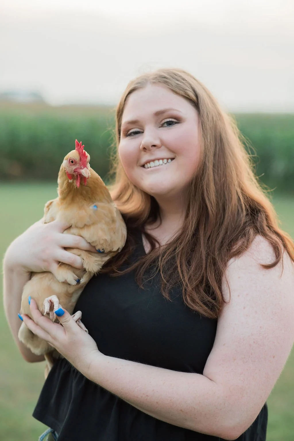 A woman with long red hair smiling and holding a tan chicken in her arms outdoors with a green field behind her.