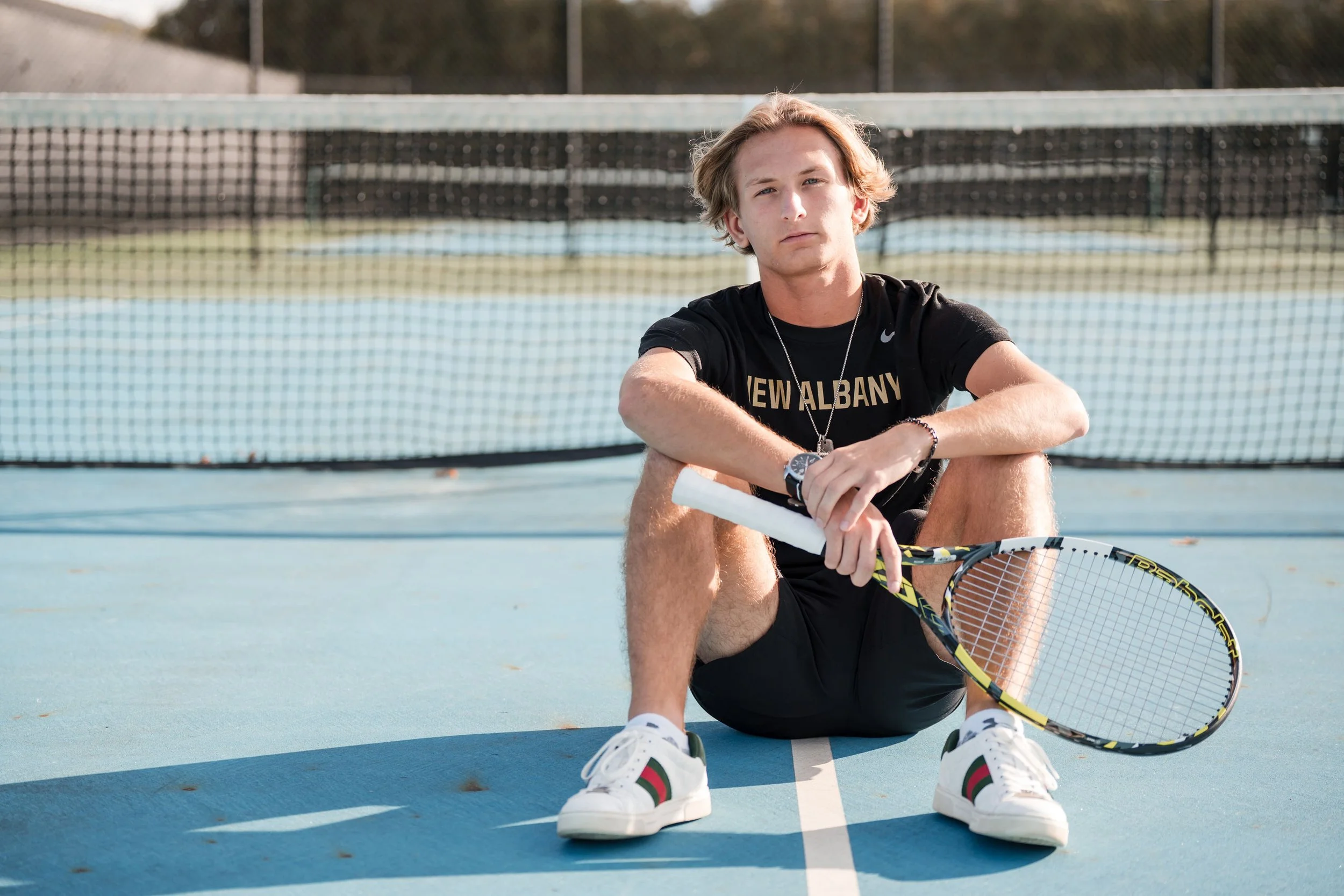 A young man sitting on an outdoor tennis court with a tennis racket and a white towel, wearing a black athletic shirt that says "New Albany", shorts, and white sneakers.