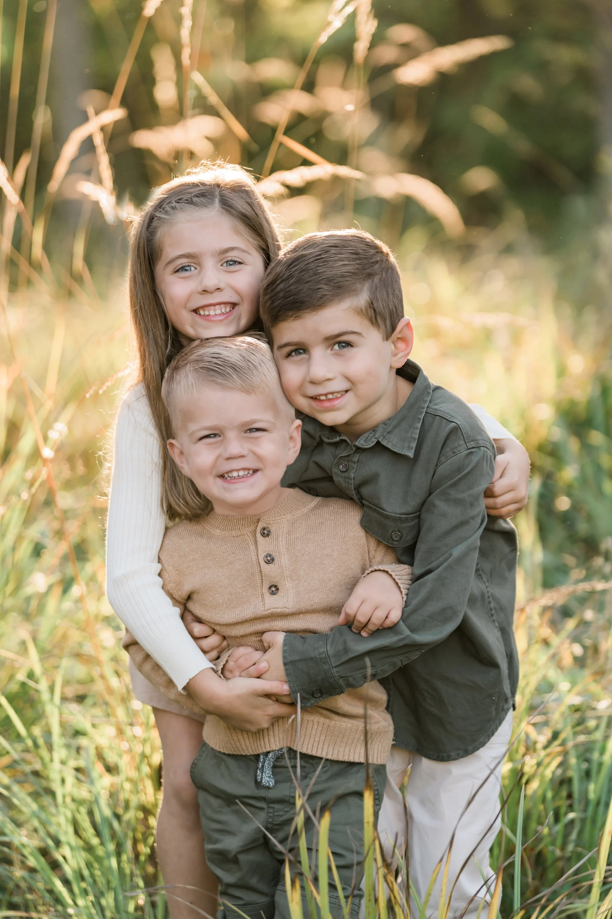 Three children hugging outdoors in a field with tall grasses during sunset.
