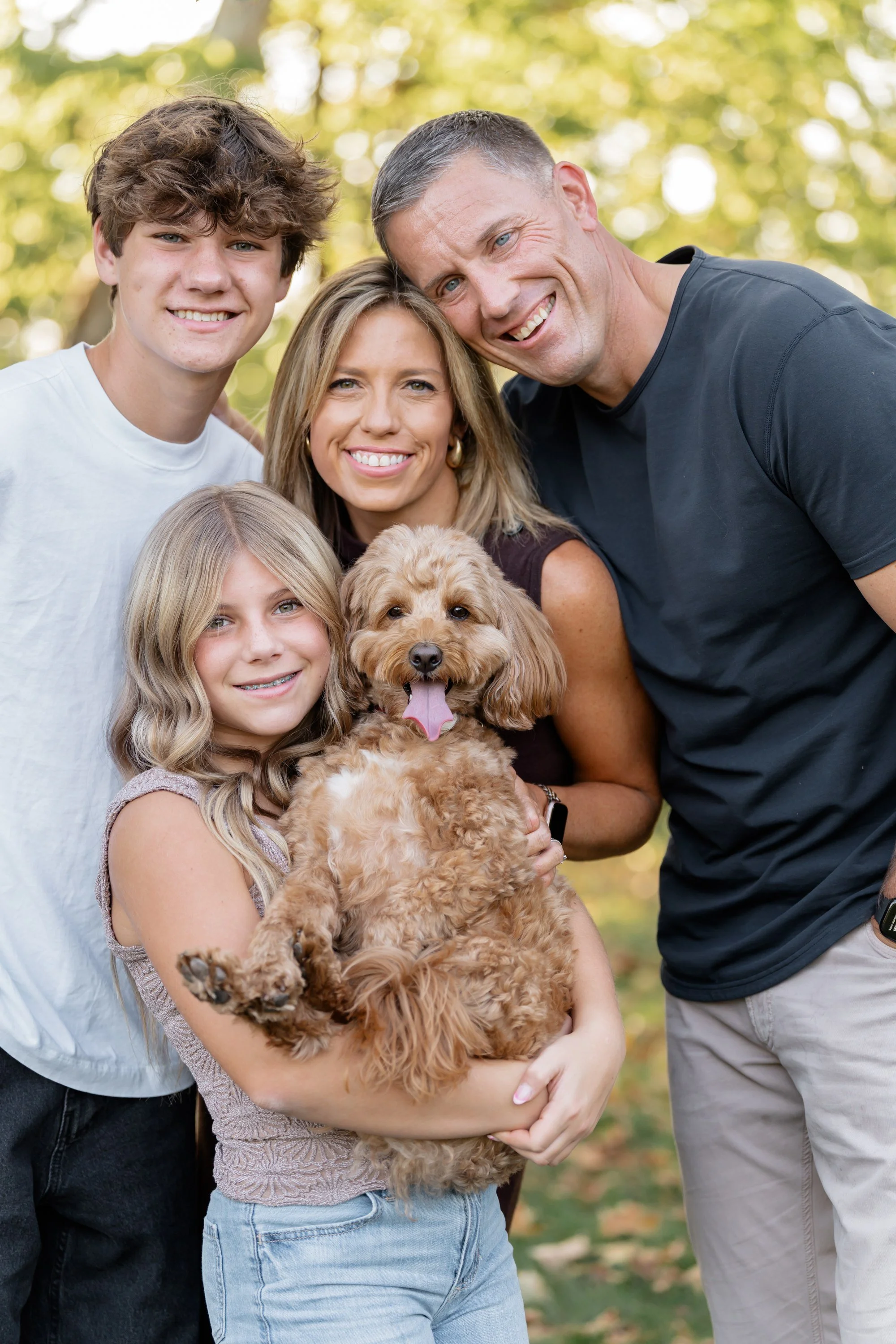 A family of four with a dog outdoors, smiling and posing for a photo during daytime, with trees and sunlight in the background.