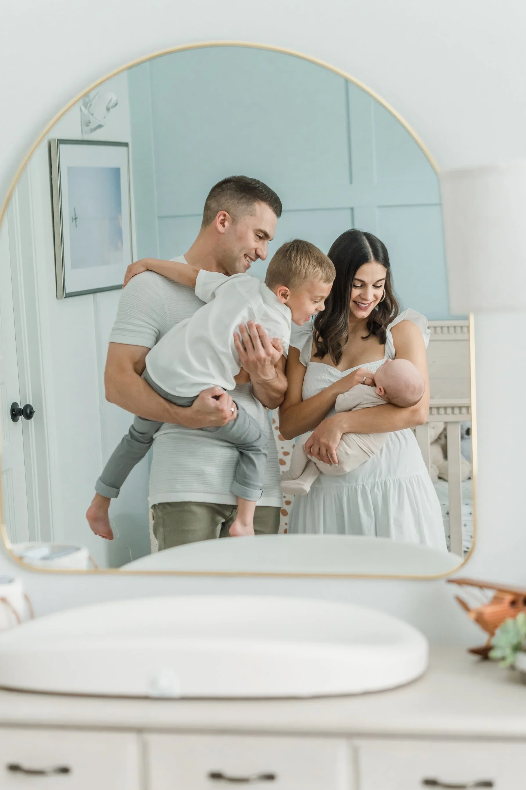 A family of four looking at a newborn baby through a mirror in a nursery, smiling and happy.