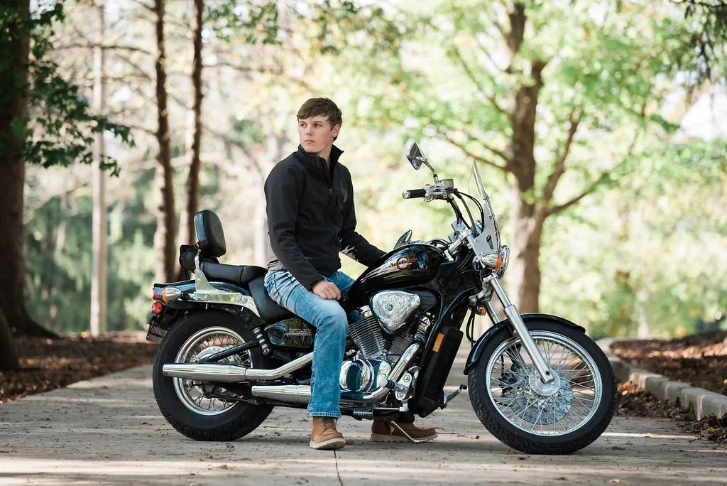A young man wearing a black jacket and jeans sitting on a black motorcycle on a paved path surrounded by trees with green leaves.