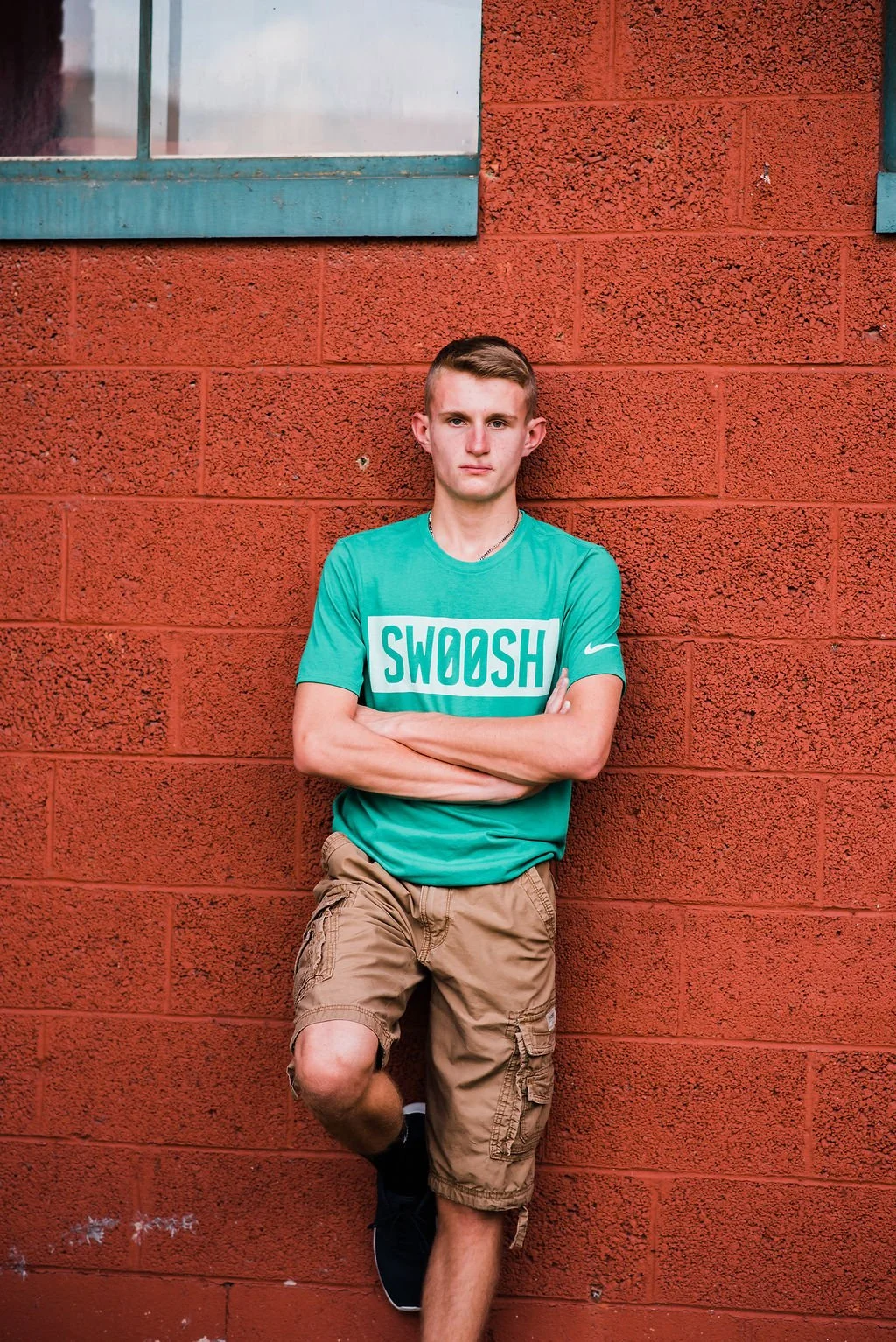A young man with short hair leaning against a red brick wall with a paint window above. He is wearing a turquoise T-shirt with the word "SWOOSH" on it, tan cargo shorts, black sneakers, and has his arms crossed.
