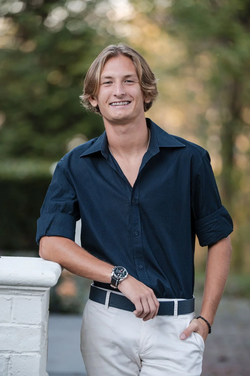 Young man with blonde hair smiling, wearing a dark blue shirt with rolled-up sleeves, light-colored pants, a watch, and a bracelet, standing outdoors near a white railing with a blurred green and yellow background.