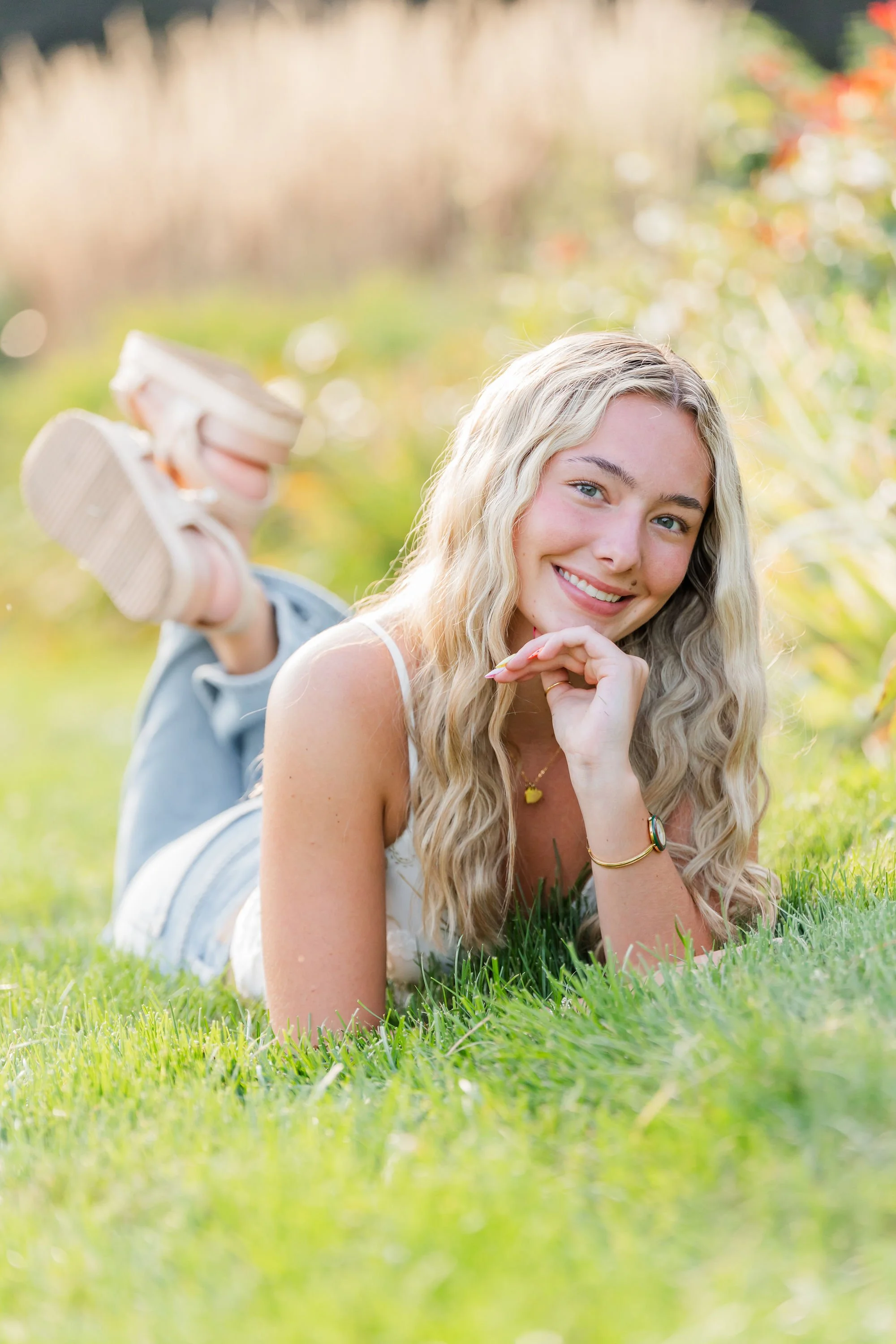 A young woman with long blonde curly hair lying on her stomach on grass outdoors, smiling at the camera with her chin resting on her hand, wearing a white sleeveless top and jewelry, with a blurred natural background.
