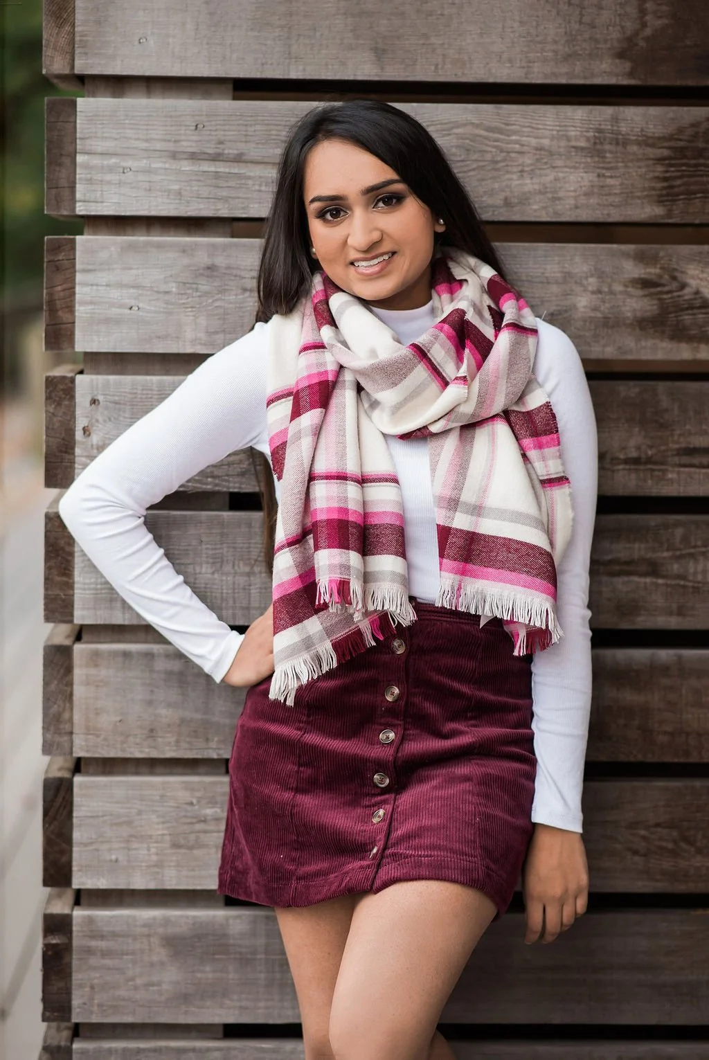 A young woman with dark hair and a friendly smile standing outdoors in front of a wooden fence. She is wearing a white long-sleeve top, a pink and white plaid scarf, and a maroon corduroy skirt.