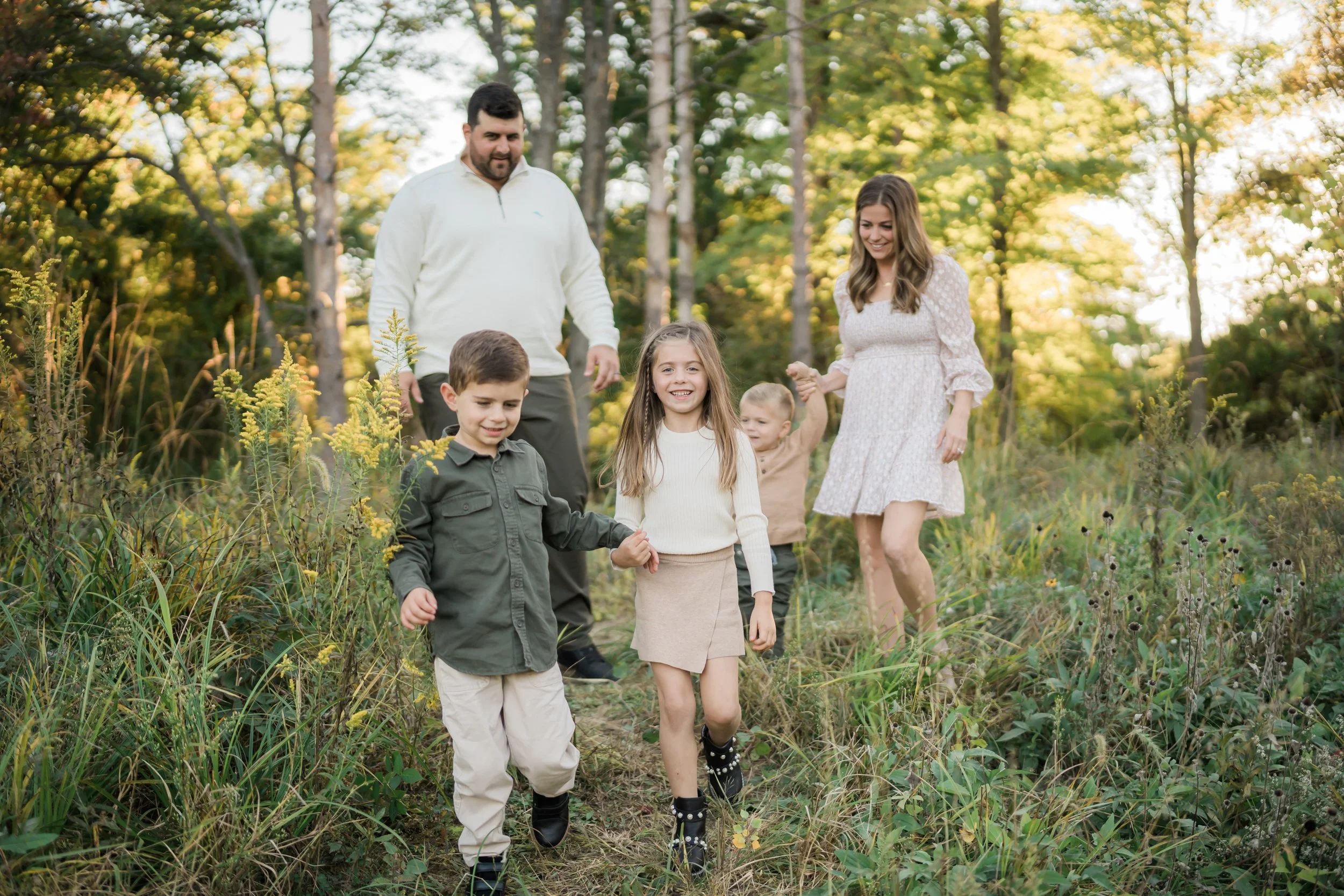 A family of six walking through a grassy forest trail during fall, smiling and holding hands.