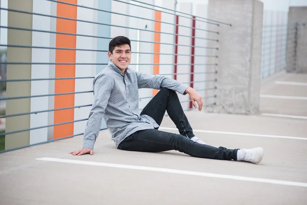 A young man sitting on a parking lot ground, leaning against a colorful fence, smiling at the camera.