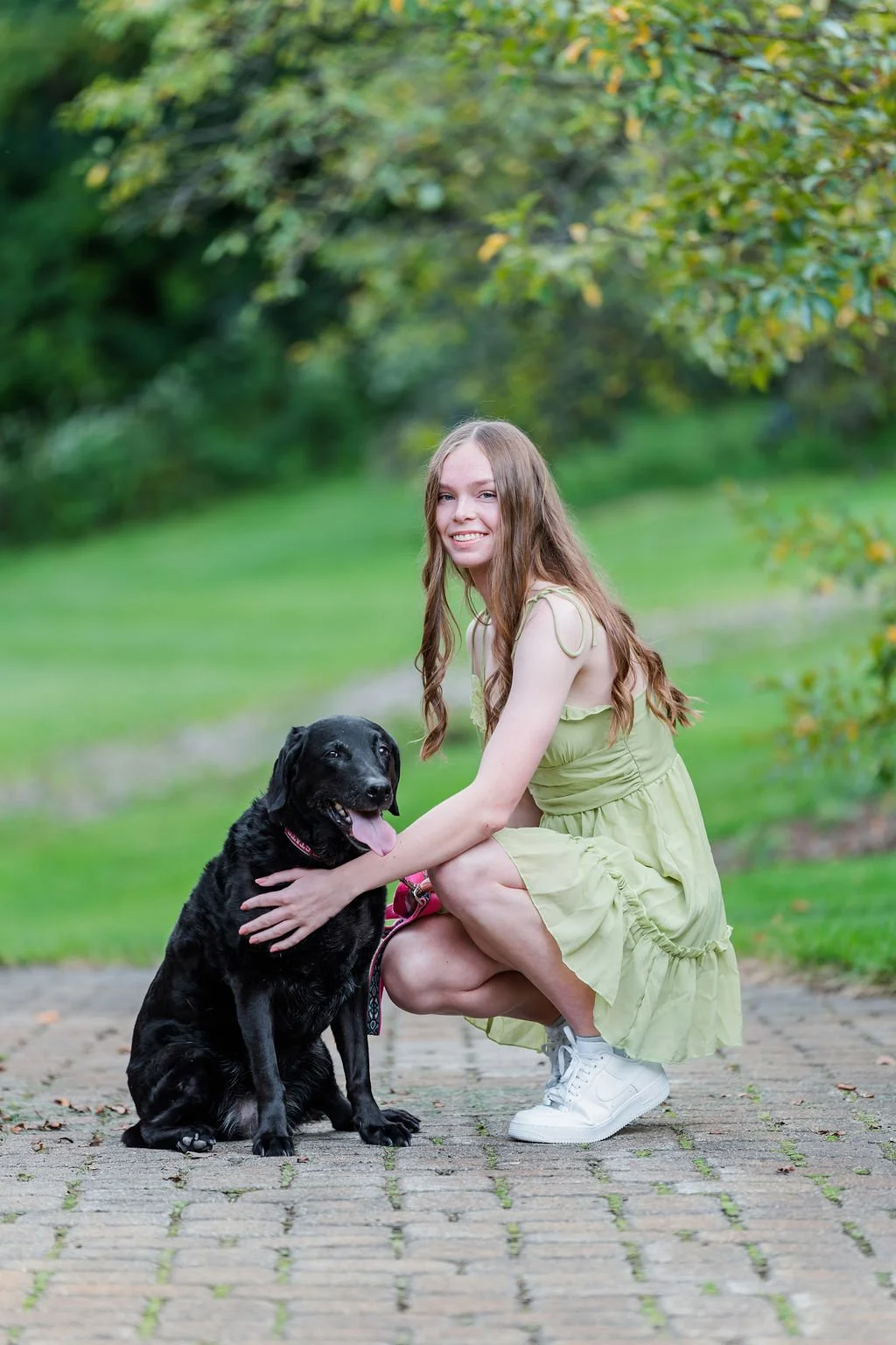 A young woman with long, wavy hair, wearing a light green dress and white sneakers, kneels on a brick pathway. She is smiling and hugging a black Labrador Retriever with a pink collar, sitting on the pathway. The background features lush green grass 