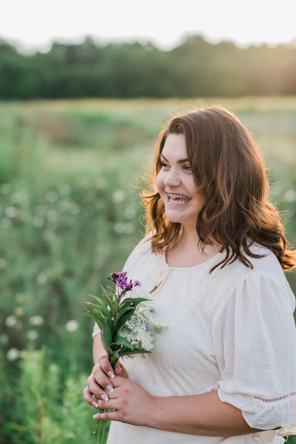 A young woman with curly brown hair and braces standing outdoors in a field during sunset, holding a small bouquet of purple and white flowers, smiling.