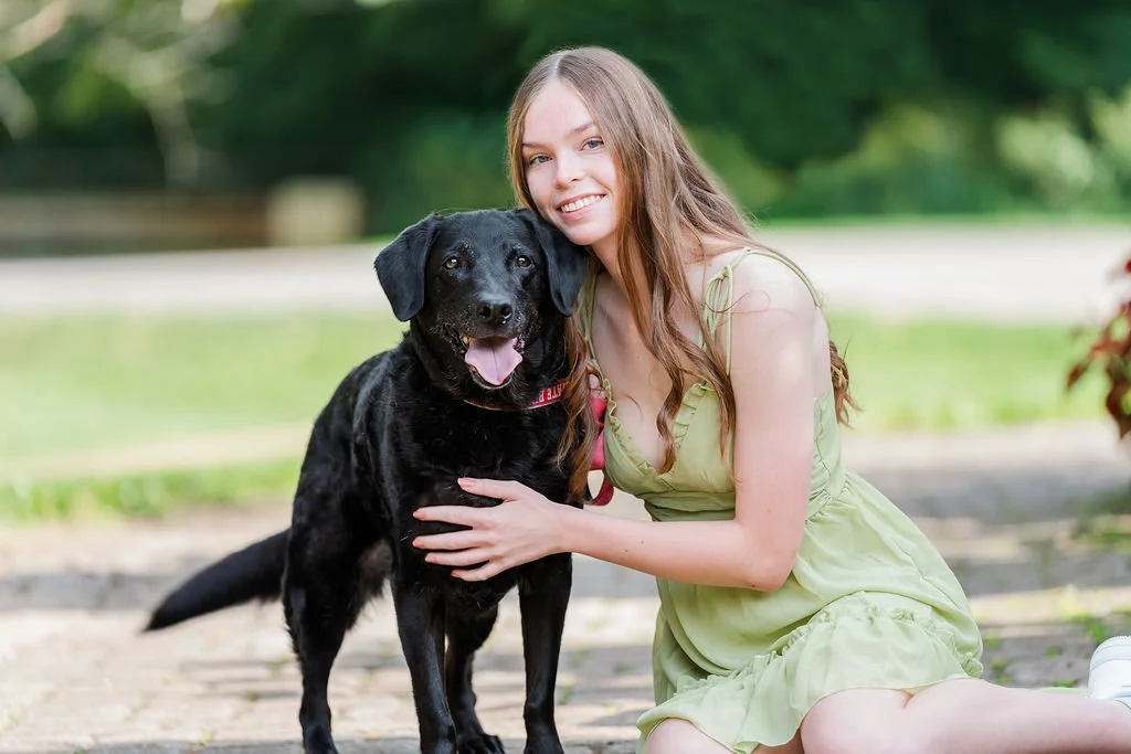 A young woman with long brown hair wearing a light green dress kneels beside a black dog with brown markings in an outdoor park setting.