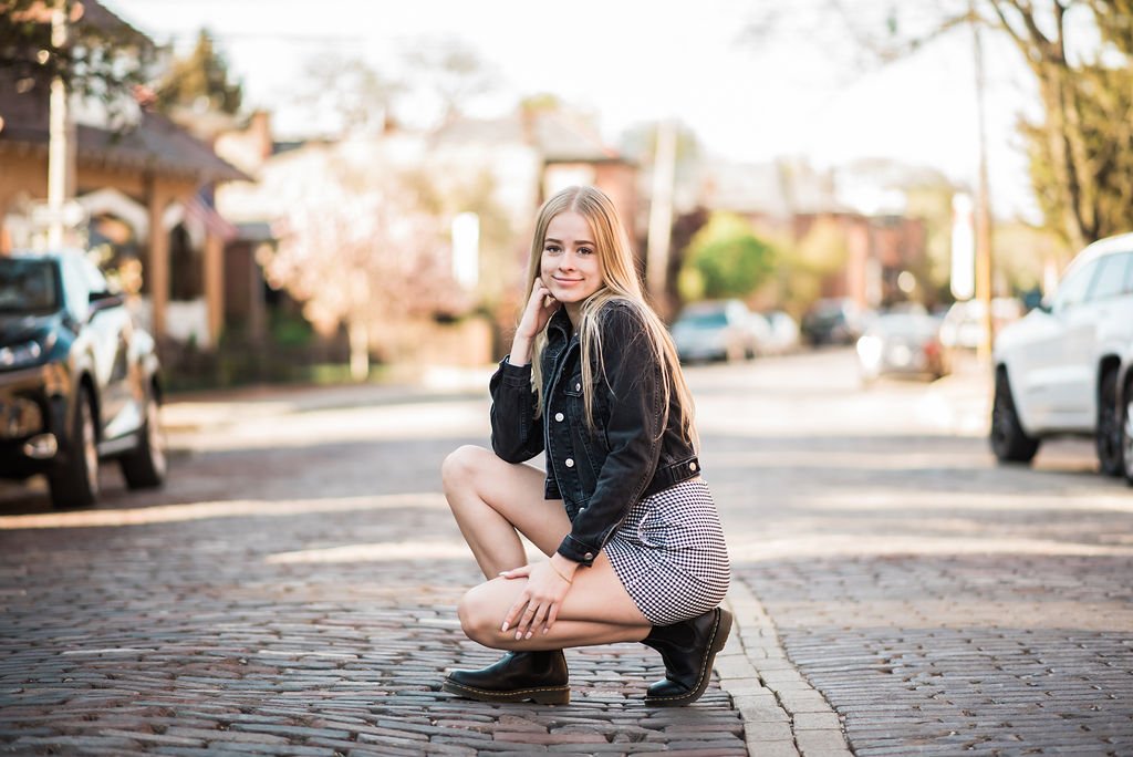 Young woman squatting on cobblestone street, smiling, wearing black jacket, checkered shorts, and black boots, in a suburban neighborhood with parked cars and trees.