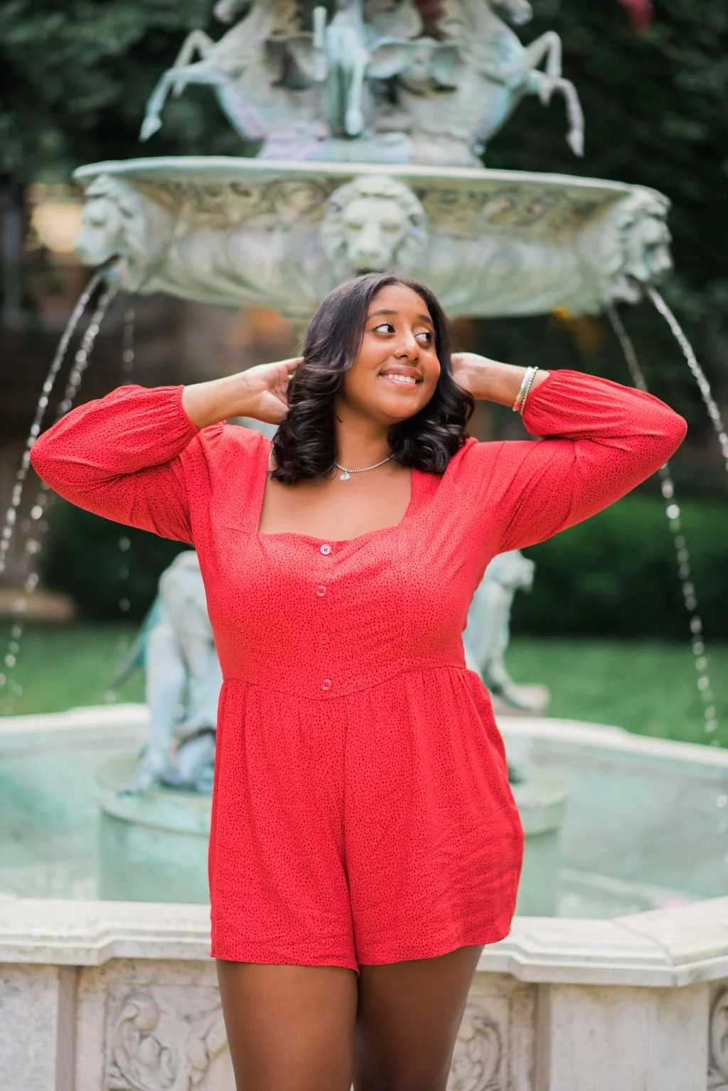 A woman in a red dress standing in front of a decorative fountain outdoors.