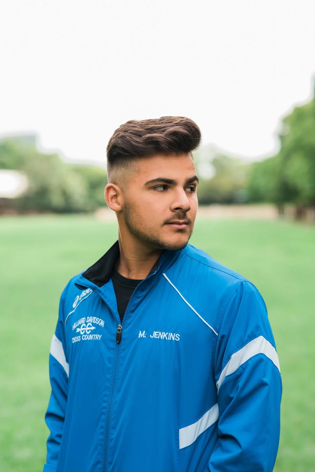 Young man in a blue athletic jacket with white accents and the name M. Jenkins, standing outdoors on a grassy field with trees in the background.