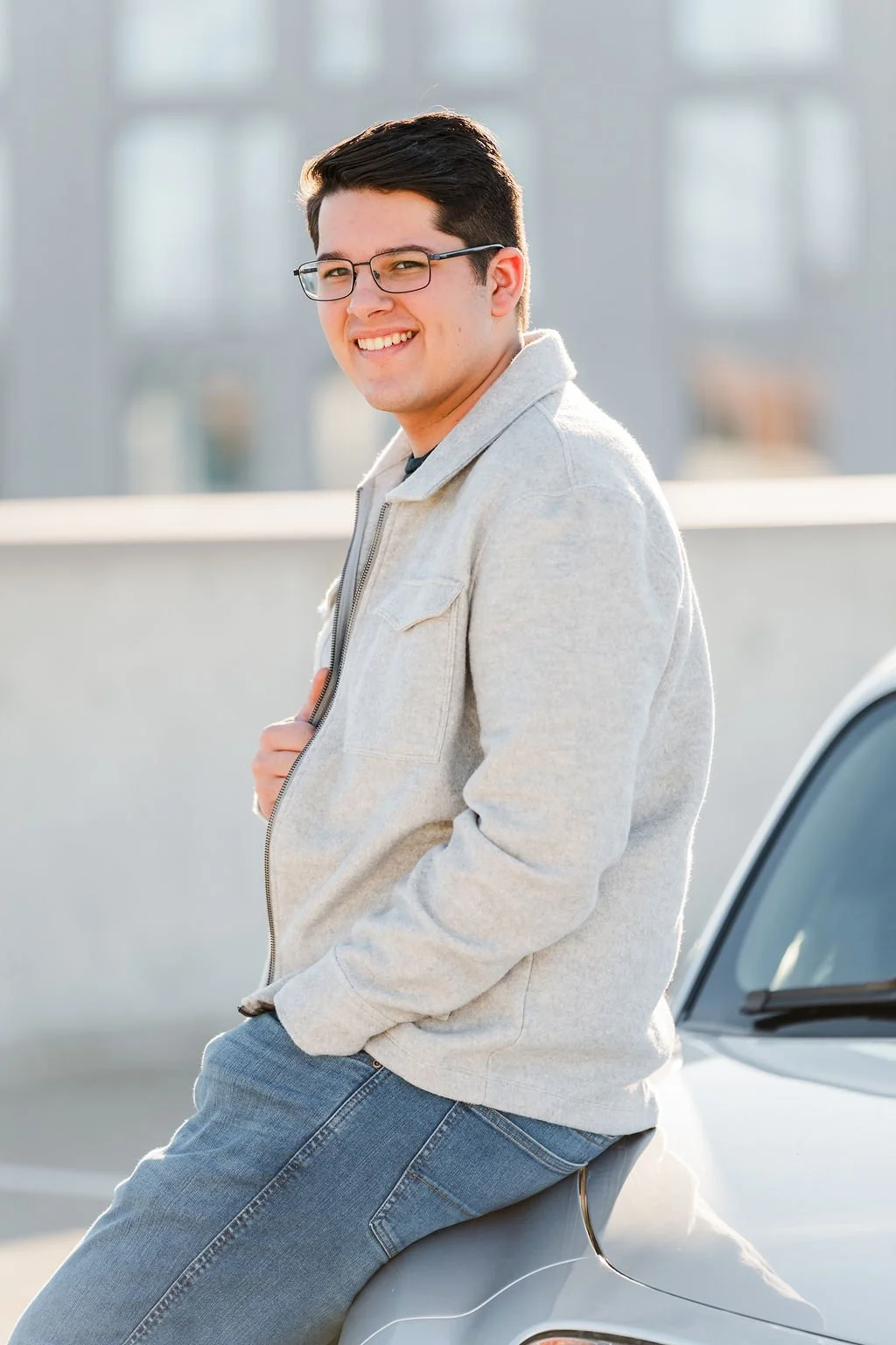 A young man with dark hair, glasses, and a light gray jacket, smiling and leaning against a silver car on a rooftop parking lot, with buildings in the background.