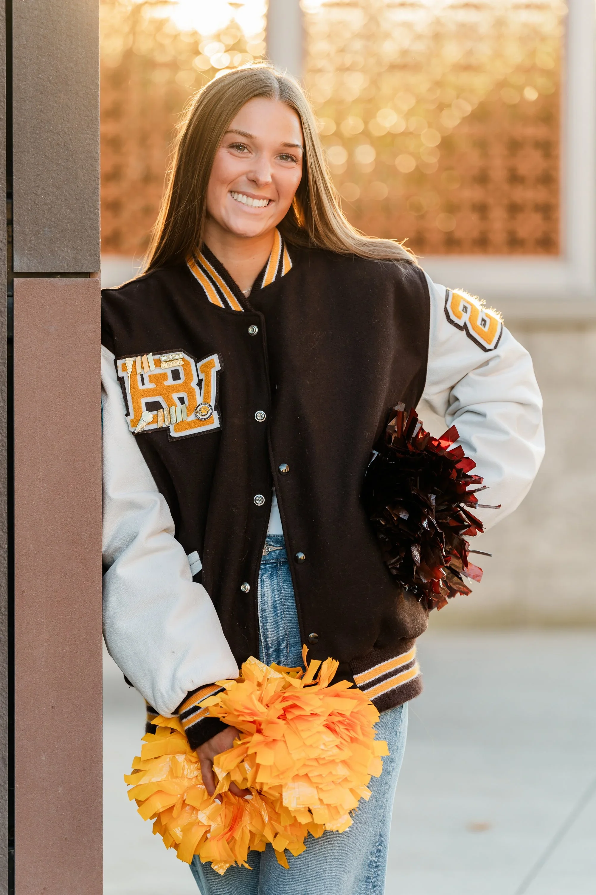 A young woman smiling outdoors at sunset, wearing a black and white varsity jacket with orange and yellow patches, holding yellow pom-poms, with a blurred background of trees.