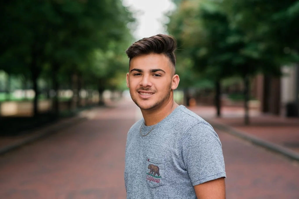 Young man standing outdoors on a city street surrounded by trees, smiling and winking at the camera.
