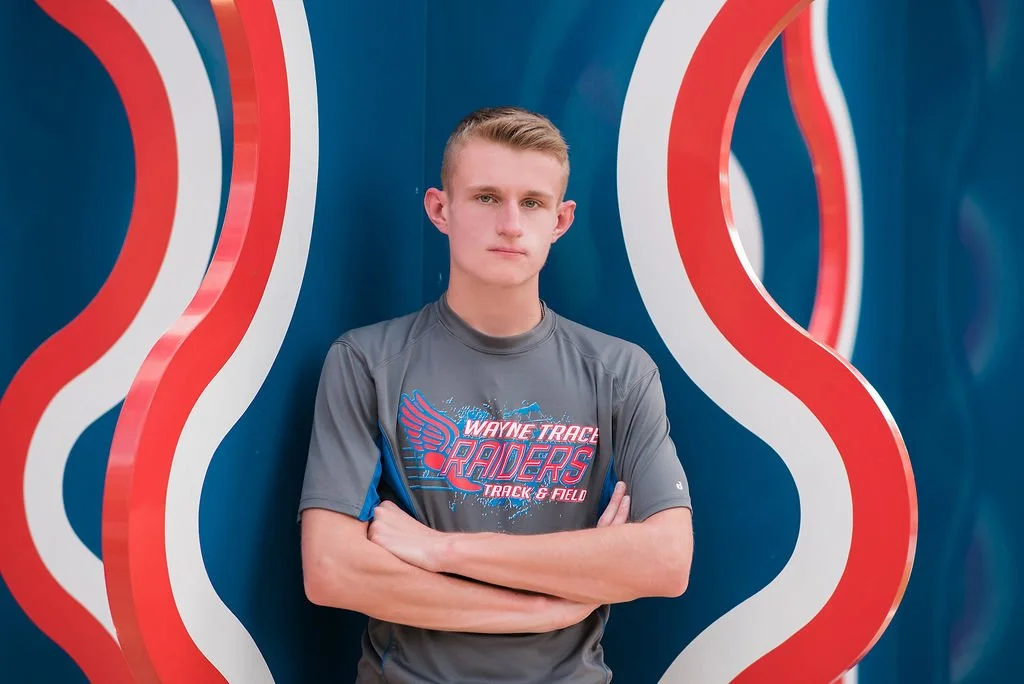 A teenage boy with short blonde hair standing with arms crossed in front of a colorful, abstract background featuring curved red, white, and blue lines. He is wearing a gray sports t-shirt with a logo for Wayne Trace Raiders Track & Field.