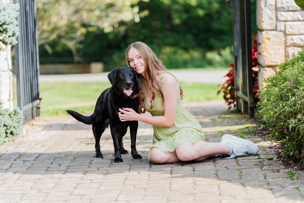 A young woman in a light green dress kneeling on a brick pathway, hugging a black Labrador Retriever dog. They are outdoors, next to a stone wall and gate, with greenery in the background.