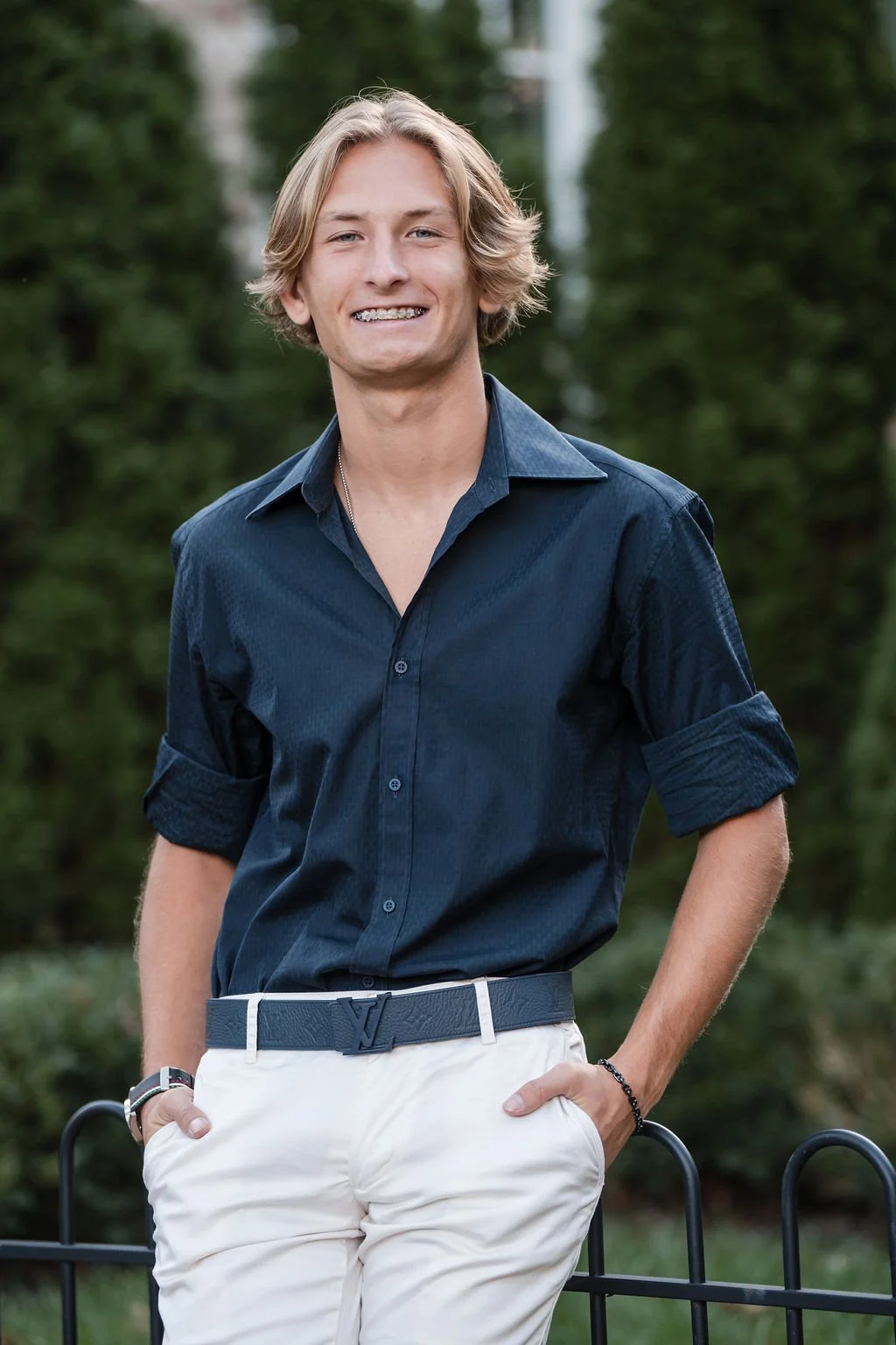 A young man with shoulder-length blonde hair smiling outdoors, wearing a dark blue button-up shirt with rolled-up sleeves and white pants with a dark belt.