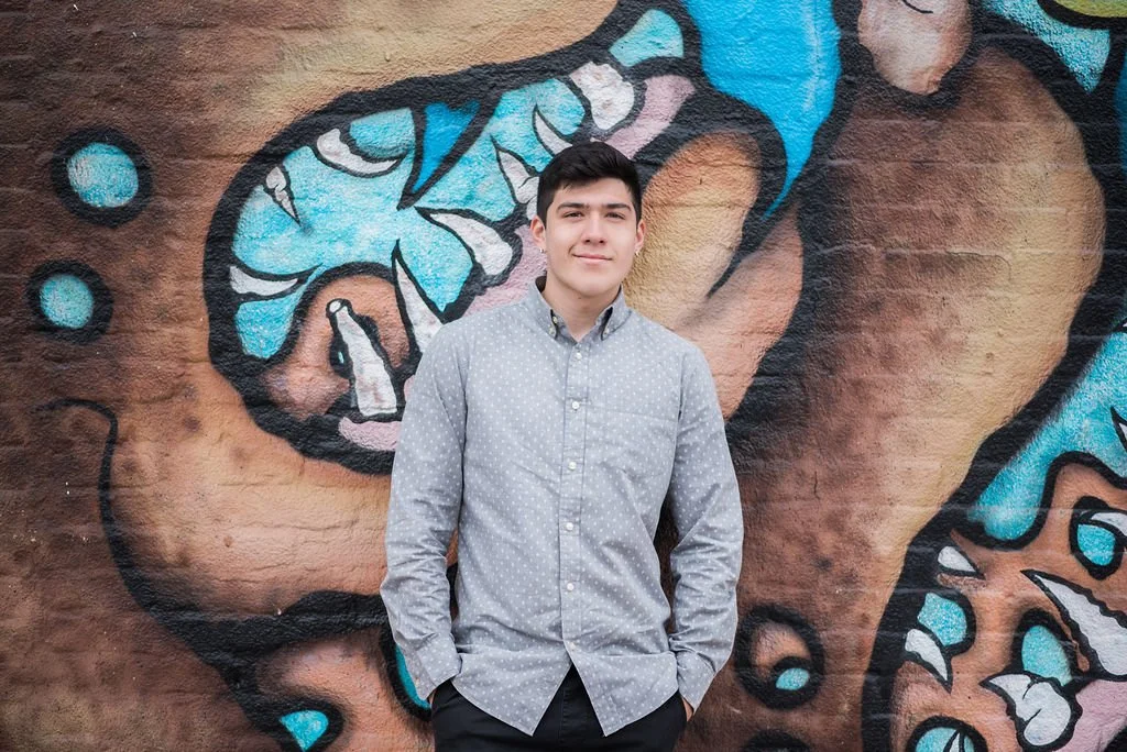 A young man in a light gray, polka-dotted button-up shirt standing in front of a colorful graffiti mural on a brick wall.
