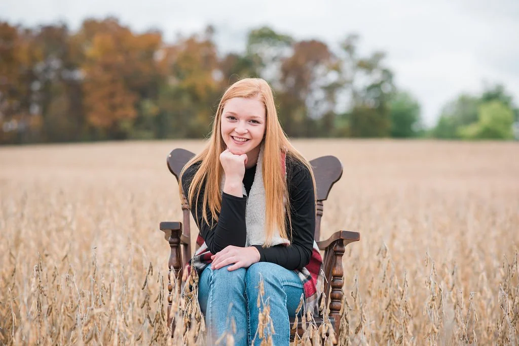 A young woman with long red hair sitting on an outdoor wooden chair in a wheat field, smiling and resting her chin on her hand, with trees in the background.