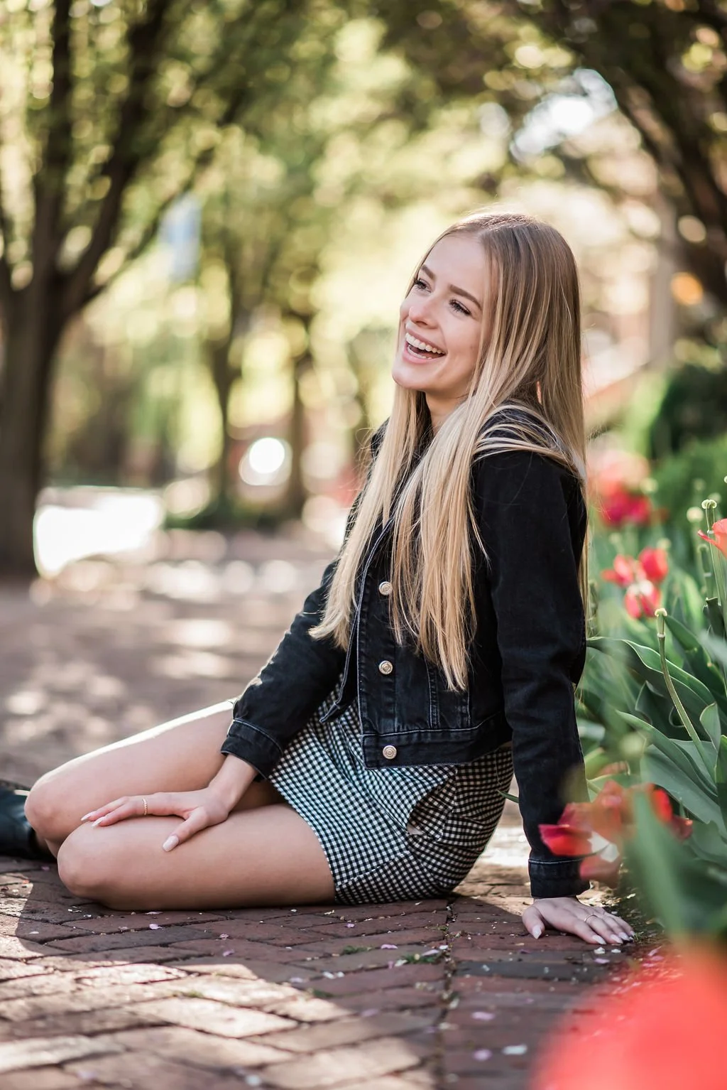A young woman with long blonde hair sitting on a brick path in a park, smiling and looking to the side, surrounded by green trees and pink flowers.