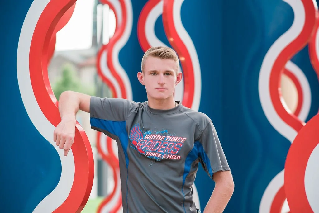 Young man in a grey and blue track shirt posing against a colorful, wavy background at an outdoor event.