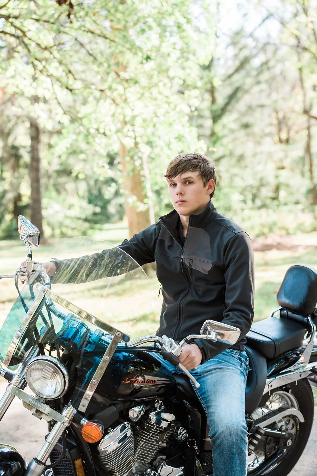 A young man in a black jacket sitting on a black Honda Shadow motorcycle outdoors in a wooded area.