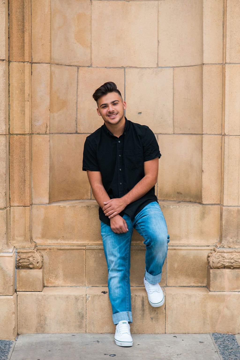A young man with dark hair dressed in a black short-sleeve shirt, blue jeans, and white shoes, sitting casually against a beige stone wall outside.