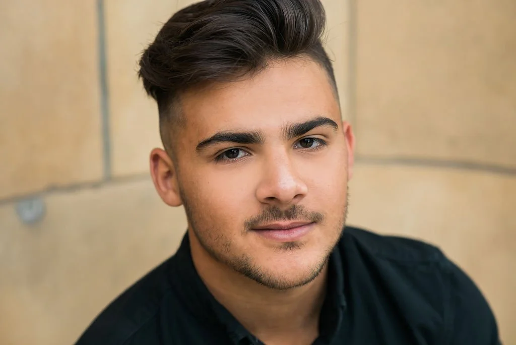 Close-up of a young man with styled dark hair, light facial hair, and wearing a black shirt, against a beige background.
