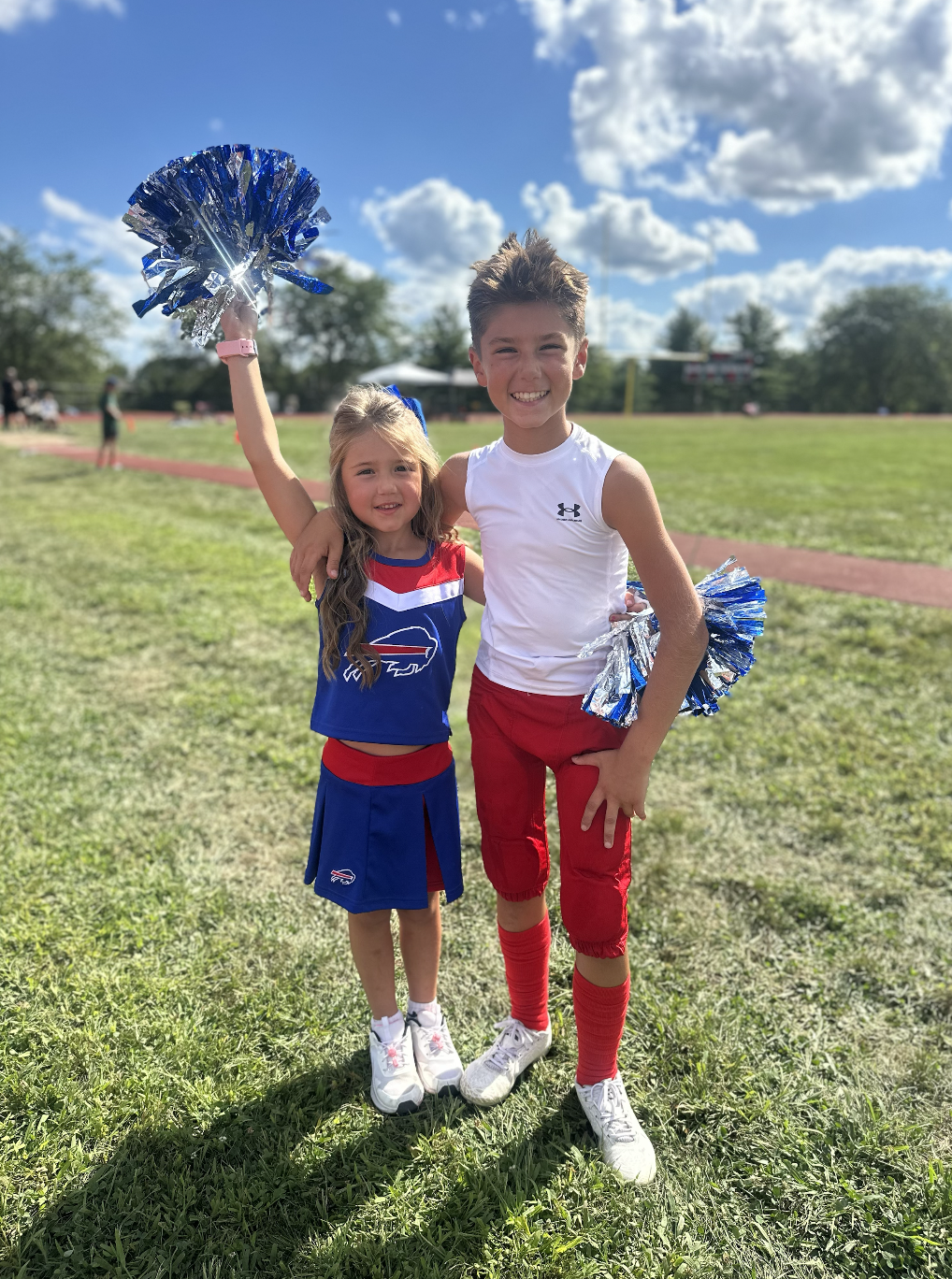 Two children, a girl and a boy, standing on a grassy field at a sports event. The girl is wearing a blue cheerleading uniform with a buffalo logo, and the boy is in a white sleeveless shirt and red athletic pants. They are smiling, holding blue and silver pom-poms, and have their arms around each other. The sky is partly cloudy.