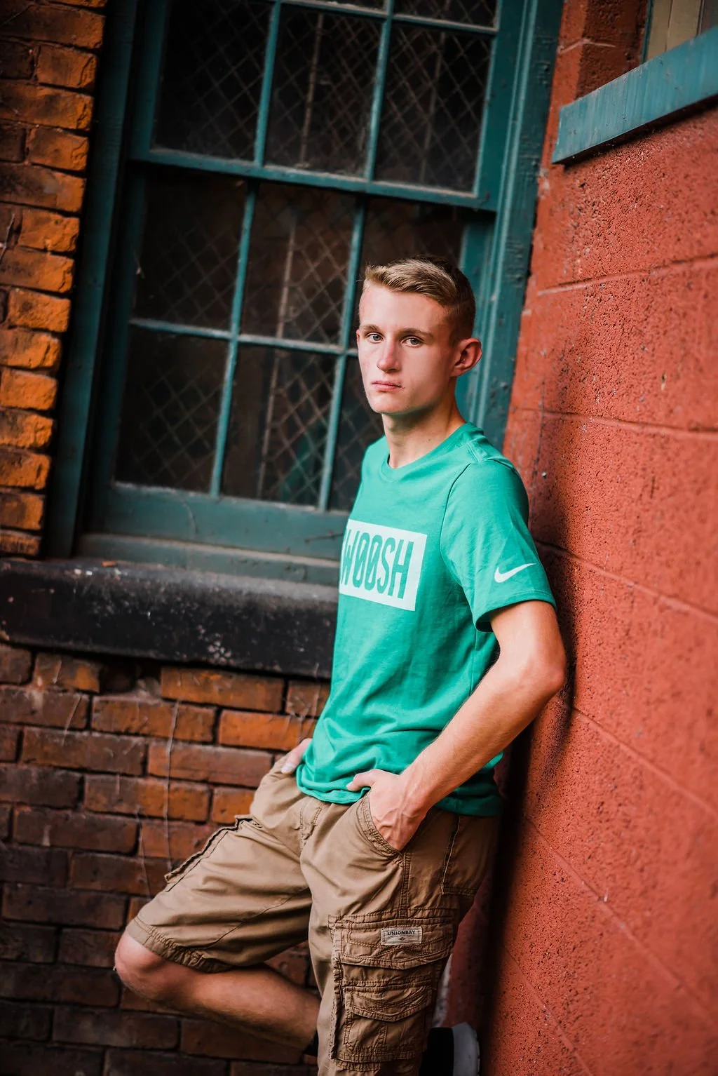 A young man with short hair wearing a teal t-shirt and beige cargo shorts leaning against a red brick wall next to a window with blue trim.