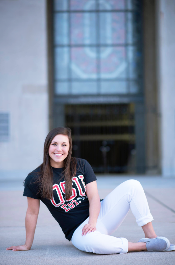 A young woman with long brown hair sits on the ground outside a building with a large glass window behind her. She is smiling and wearing a black T-shirt with red and white lettering and white pants.