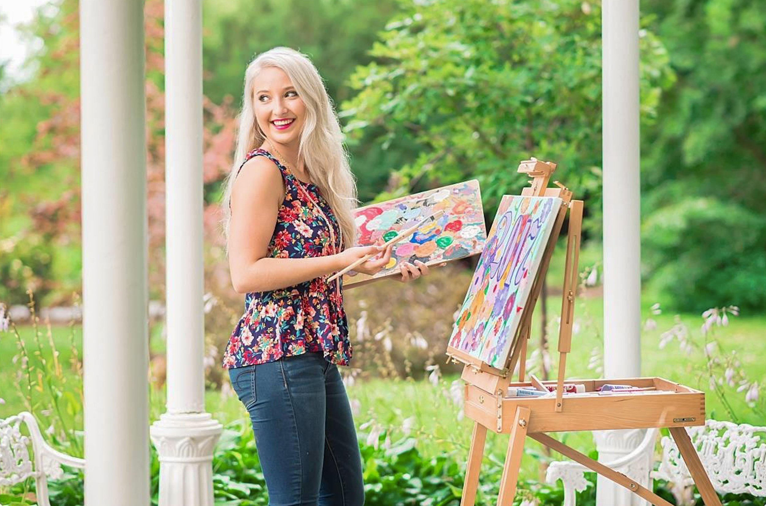 A woman with long blonde hair painting outdoors on a sunny day, standing beside an easel with colorful abstract art, in a garden with trees and flowers.