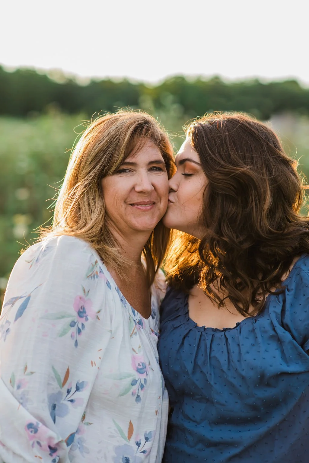 A woman with light brown hair in a white floral blouse is being kissed on the cheek by a woman with dark brown hair in a blue dress during sunset outside.