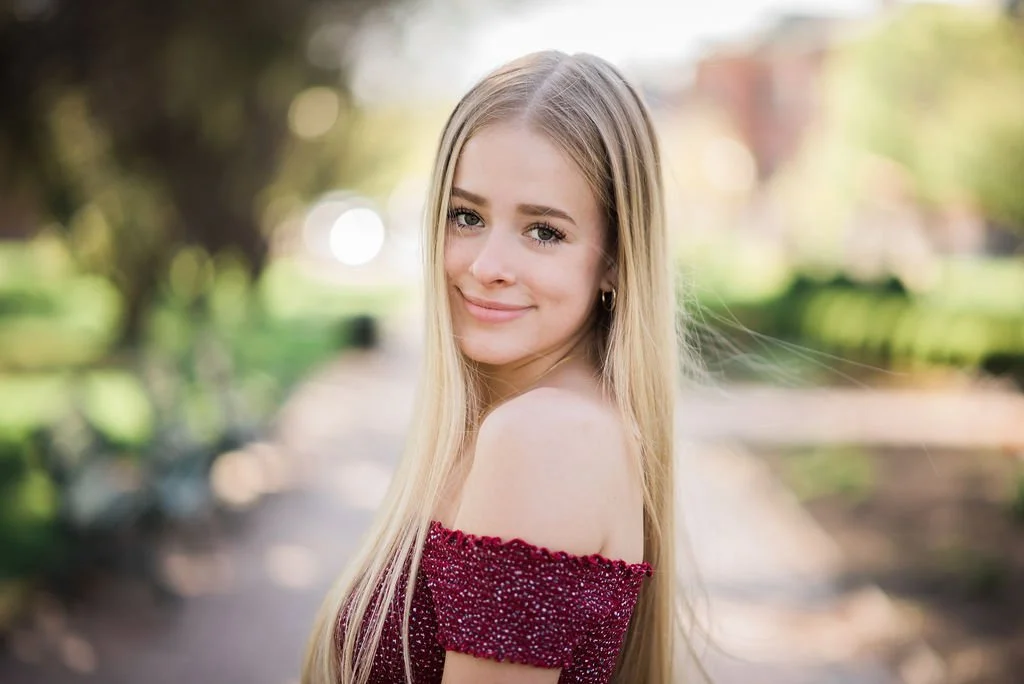 A young woman with long blonde hair smiling outdoors in a park with trees and greenery in the background, wearing a red off-the-shoulder top.