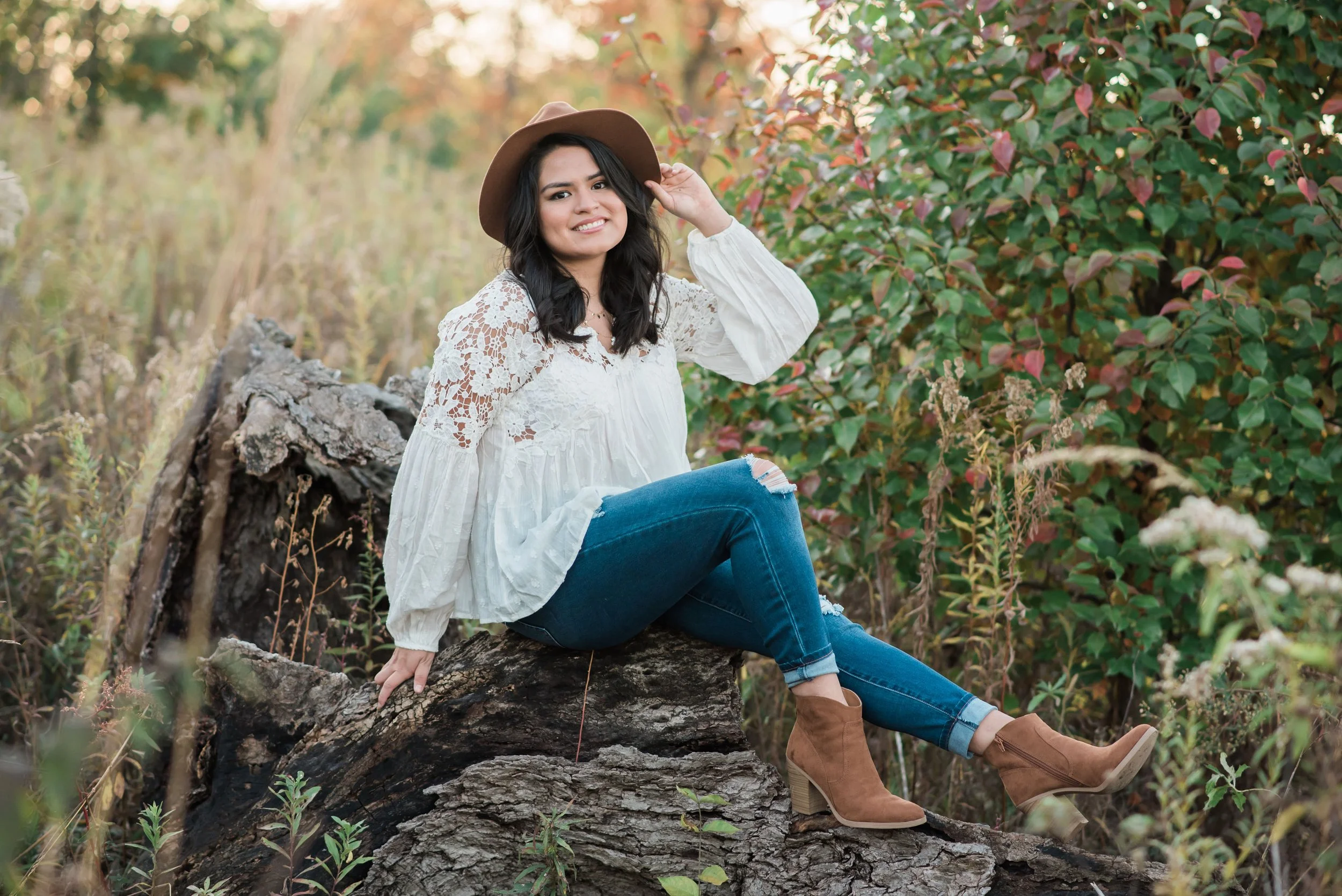 A woman sitting on a fallen tree trunk in a forest, wearing a white lace blouse, ripped blue jeans, ankle boots, and a wide-brimmed hat, smiling and looking at the camera.