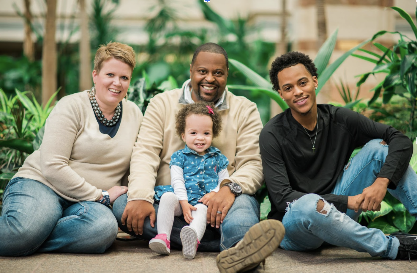 Family of five sitting on the ground indoors, surrounded by lush green plants.
