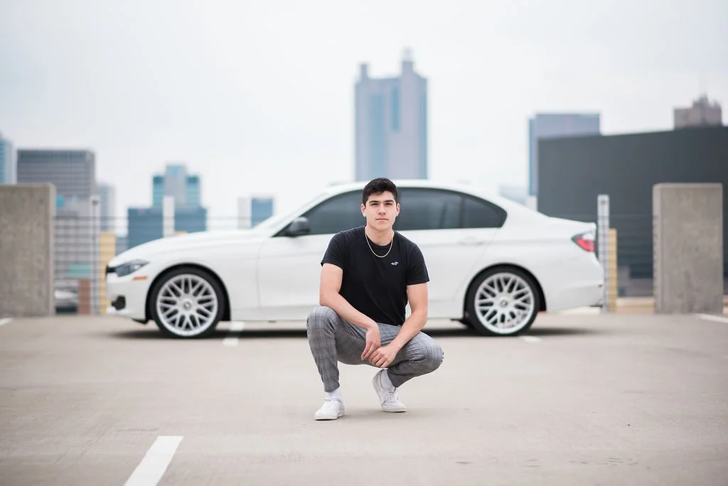 A young man squatting in front of a white car on a rooftop parking lot with city skyscrapers in the background.