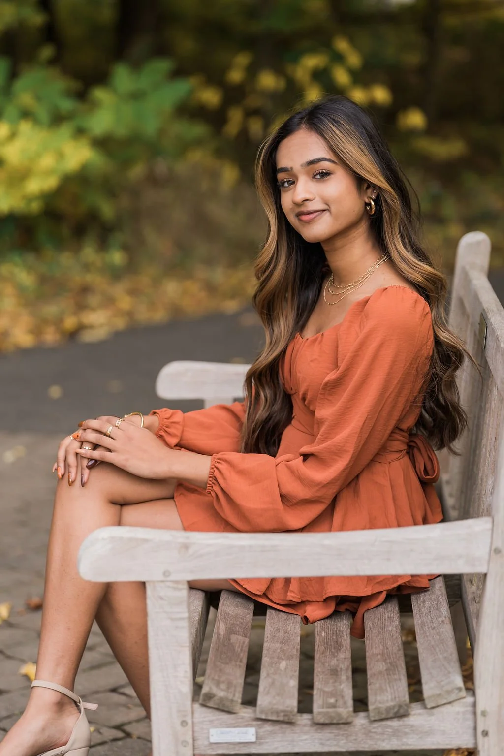 A young woman with long dark hair with highlights sits on a wooden park bench wearing an orange dress and beige heels, surrounded by fall foliage.