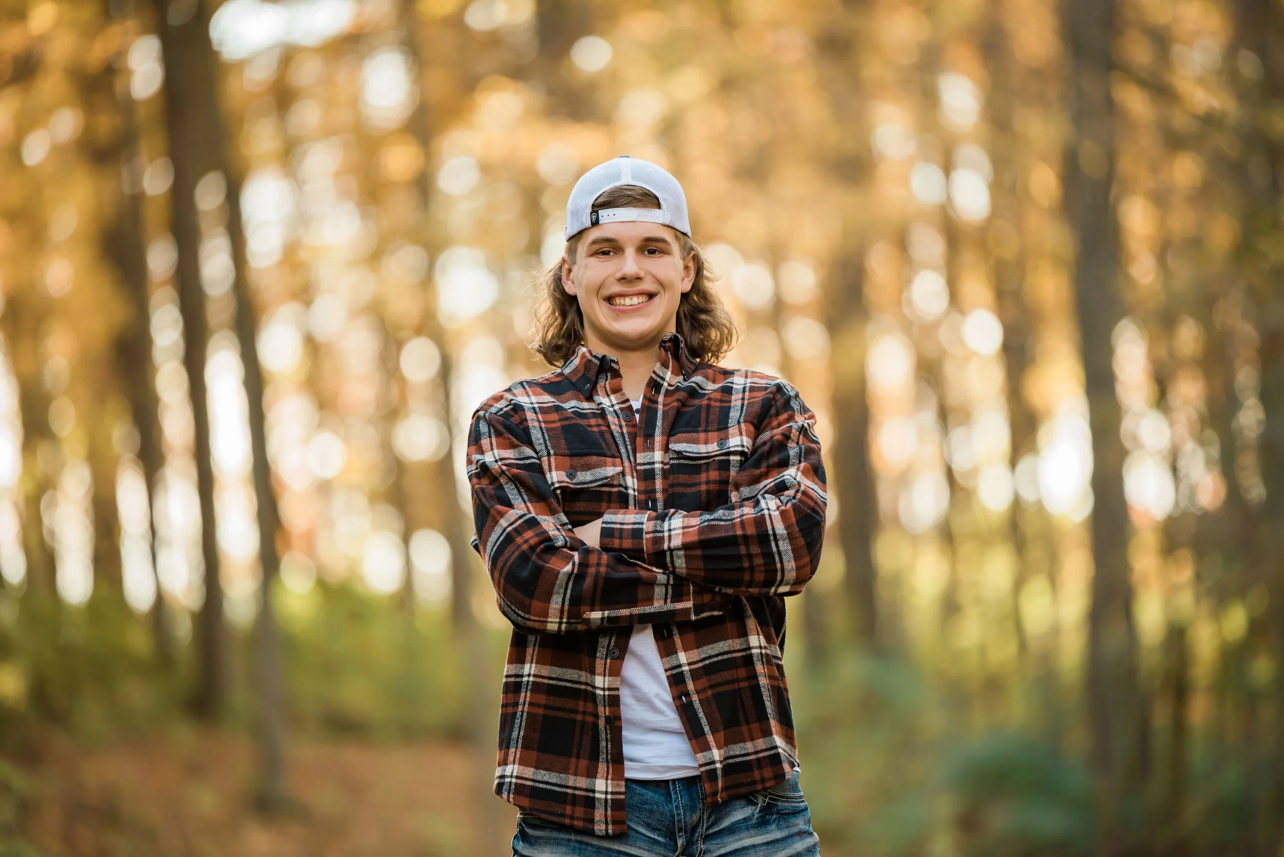 A young man with long hair, wearing a baseball cap backwards, a plaid shirt, and jeans, smiling with his arms crossed, standing outdoors in a forest during autumn.