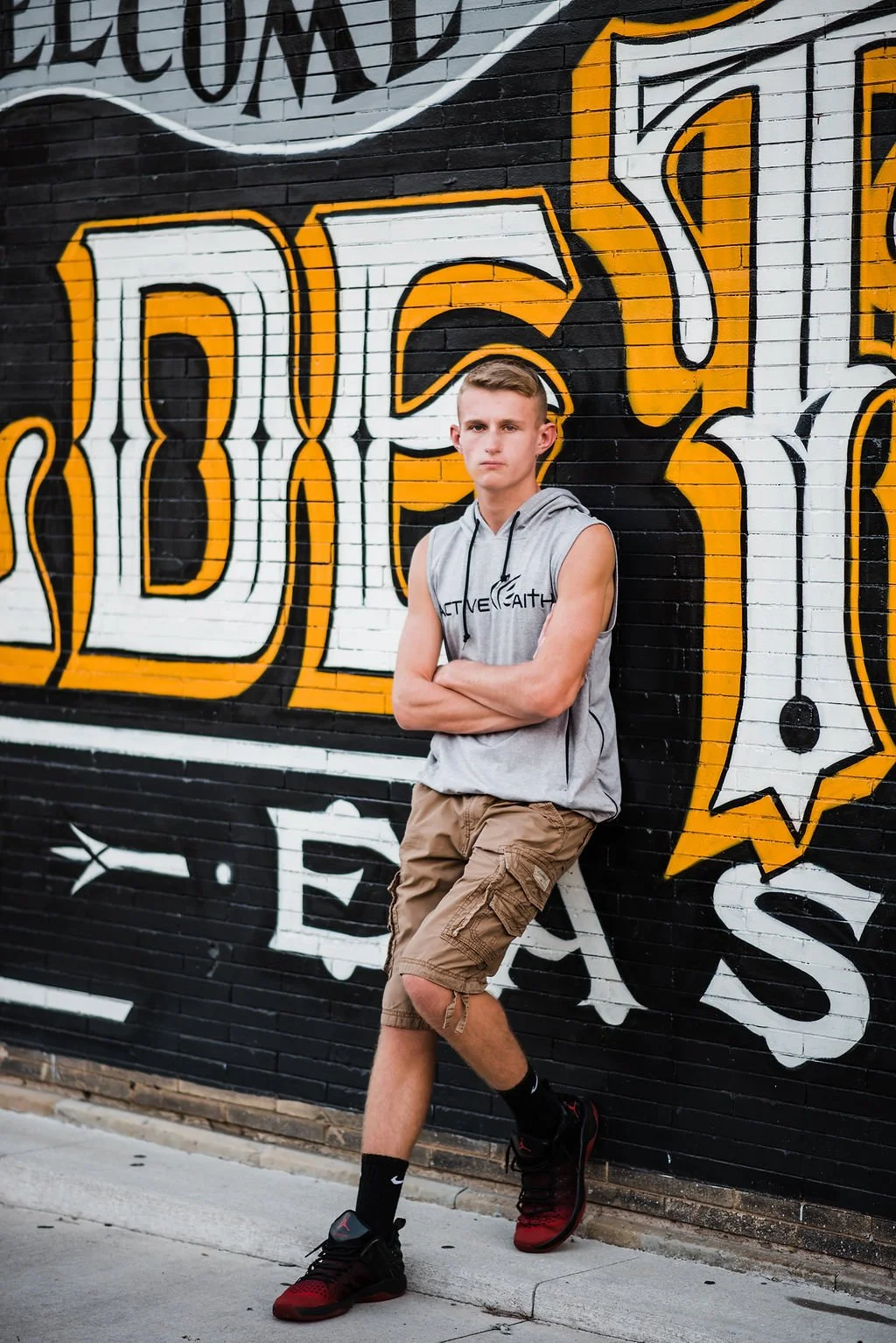 A young man with short blond hair, wearing a sleeveless hoodie with the text 'ACT ONE FAITH', brown cargo shorts, and black and red athletic shoes, stands with arms crossed against a colorful graffiti wall.