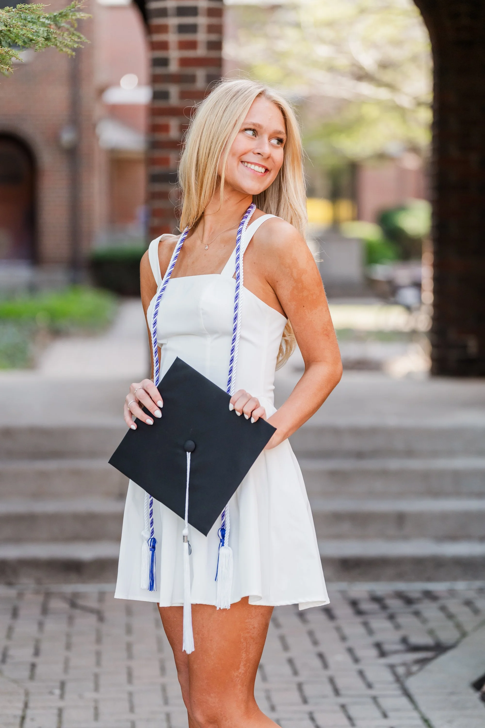 A young woman in a white dress holding a graduation cap with a blue and white tassel outdoors, smiling and looking to the side.