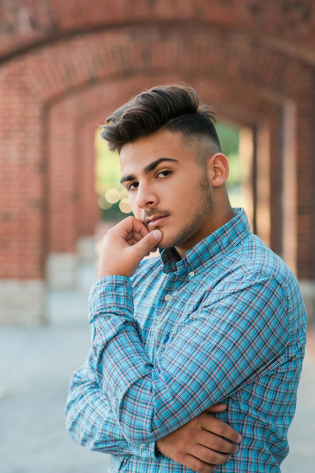 Young man with styled hair and a checked shirt standing outdoors near brick arches, posing with one hand on his chin.