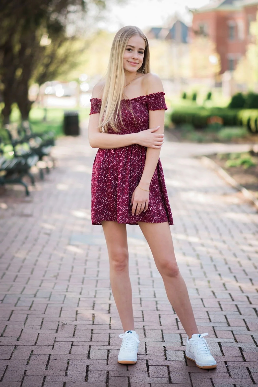 Young woman standing on a brick pathway in a park during daytime, wearing a red off-shoulder dress with white polka dots and white sneakers, with trees and buildings in the background.